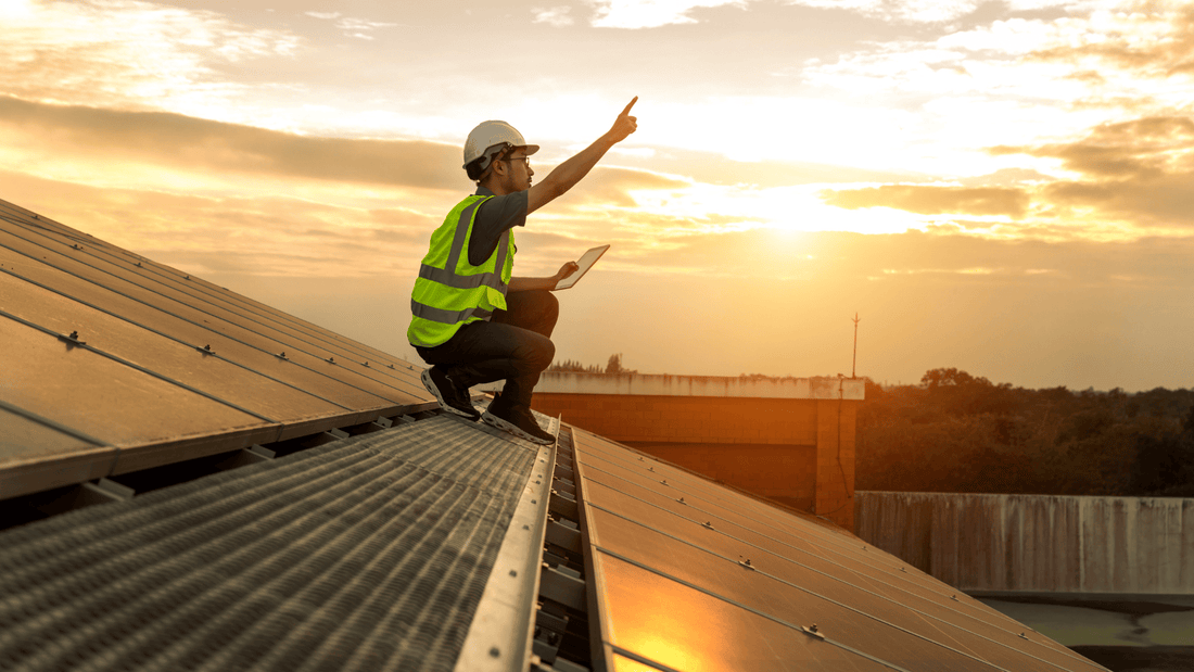 Solar installer with hard hat on roof next to solar panels; sunset in the background.